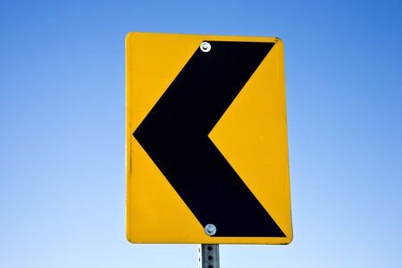 Black arrow on yellow traffic sign pointing left on blue sky, USAの写真素材