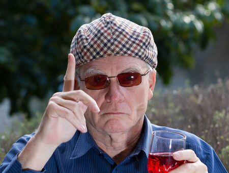 Older man in reversed flat tweed cap drinking red wine and wearing sunglassesの写真素材