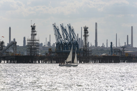 A yacht under sail passing the landscape of the Fawley Refinery on Southampton Water in Hampshire,...