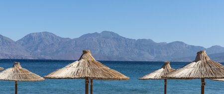 Agios Nikolaos, eastern Crete, Greece. October 2019.  Straw sun umbrellas and distance mountains at Agios Nikolaos, Eastern Crete,のeditorial素材