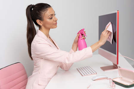 Wiltshire, England, UK. 2021.  Woman at desk using a spay bottle and cloth to clean and sanitise a computer screen.の写真素材