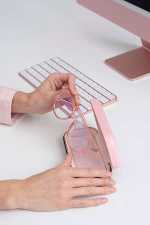 England,UK. 2021.  Womans hands removing glasses from the case with a backdrop of computer and keyboard, all pink.の写真素材