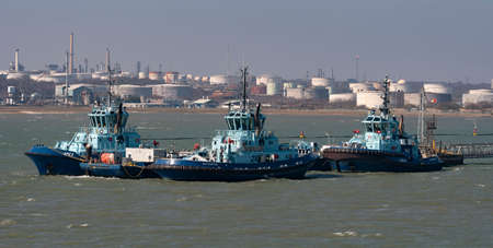 Fawley, Southampton, England, UK. 2022.  Three ocean going tugs alongside a jetty with a background of Fawley Refinery, storage tanks, chimneys and smoke stacks,の写真素材