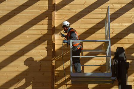 Tradesman spray painting the wall of a wooden industrial building with timber preservative