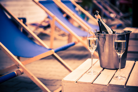 A Bottle of Champagne in Wine Cooler with Ice and Two Full Glasses on Small Wooden Pallet Table. Lodges in the Background. Summer Holiday Rest.の写真素材