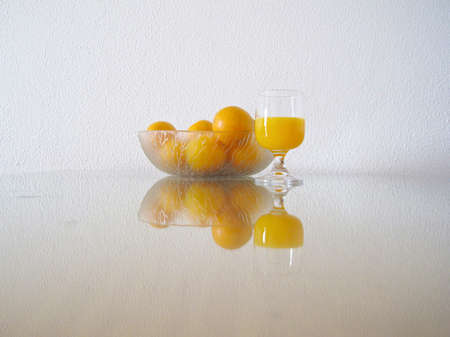 freshly squeezed orange juice and bowl of oranges reflecting off of a glass table with white wall in the background.の写真素材
