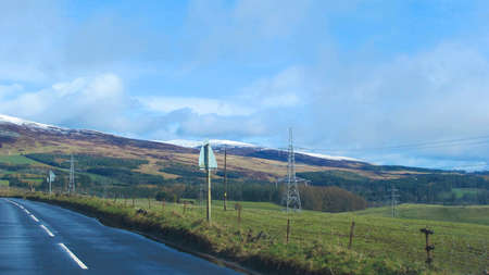 road through the hillside in scotland with electricity pylons, snow covered hills and cloudy blue sky in view.の写真素材
