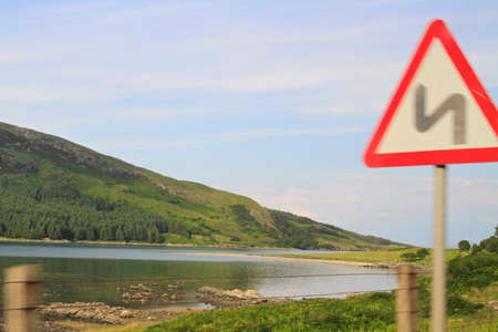 bend in the road sign with trees and hills in the background and white clouds and blue sky above.の写真素材