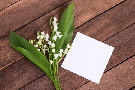 delicate white lilies of the valley with green leaves on a brown wooden background with a sheet of white paper for text.の写真素材