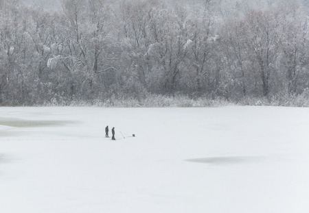 The river is covered with ice and covered with snow. Two people catch fish on winter fishing. On the opposite shore trees in the snowの写真素材