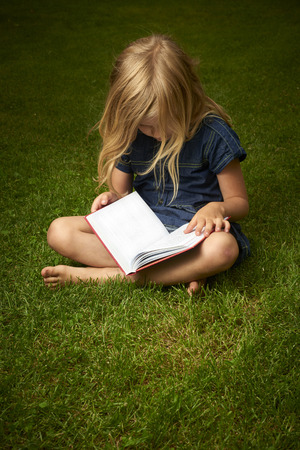 Cute Little Blond Girl Reading Book Outside on Grassの写真素材