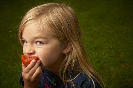Cute blond child girl eating tomato outside in the gardenの写真素材