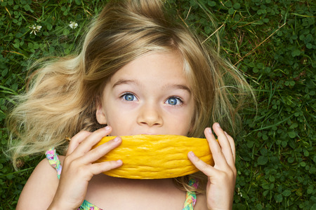 Cute happy little blond girl with melon lying on the grass in summertimeの写真素材