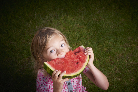 Cute little blond girl eating watermelon on the grass in summertimeの写真素材