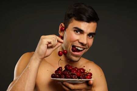 Smiling man eats red fresh cherry on gray backgroundの写真素材