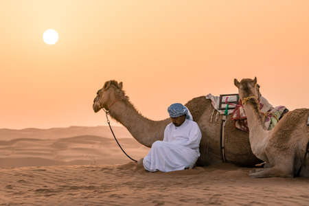 Wahiba Sands,Oman - 04.07.2018: A man in white robe and his camel sit on the desert sand. Early morning in Wahiba Sands, Oman. Nomadic life.のeditorial素材