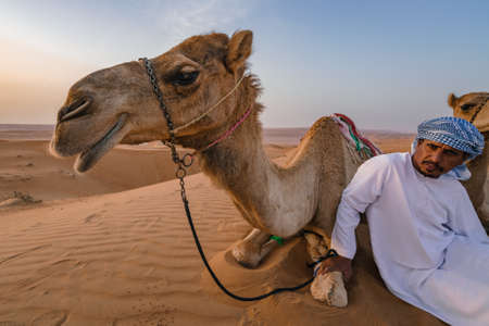 Wahiba Sands,Oman - 04.07.2018: A man in white robe and his camel sit on the desert sand. Early morning in Wahiba Sands, Oman. Nomadic life.のeditorial素材
