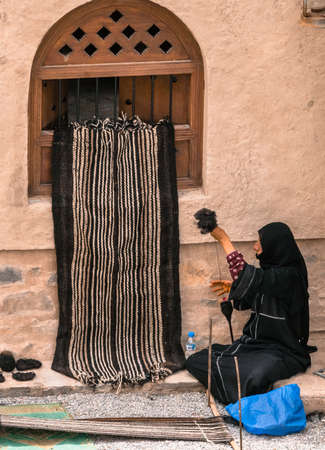 Nizwa, Oman - 04.06.2018: Woman in tradiitonal black abaya makes a fabric in a traditional way. Traditional work in medieval arabian fort of Nizwa, Oman.のeditorial素材