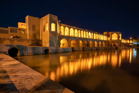 14.04.2019 - Isfahan, Iran: Famous Khajoo Bridge in Isfahan, Iran, during night. Old stone bridge lit with yellow and orange lights reflects in water. Ancient Persia.のeditorial素材