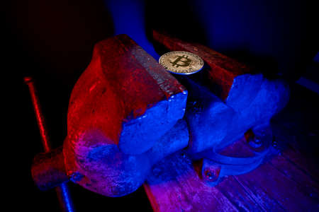 Bitcoin mounted in a steel vise in red and blue light. Keeping Bitcoin safe in crypto wallet. Pressure on crypto currency. Crypto currency restrictions. Investing instruments.の写真素材