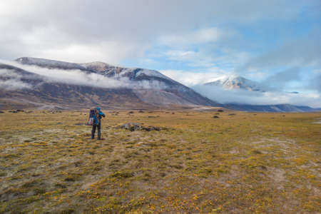 Hiker walking through remote arctic valley on a partly cloudy summer day. Dramatic arctic landscape of Akshayuk Pass, Baffin Island, Canada. Grass and moss in autumn colors.の写真素材