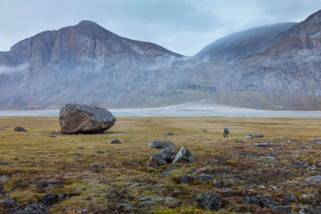 Hiker dwarfed by a big rock in remote arctic valley on a cloudy, rainy day. Dramatic arctic landscape of Akshayuk Pass, Baffin Island, Canada. Grass and moss in autumn colors.の写真素材