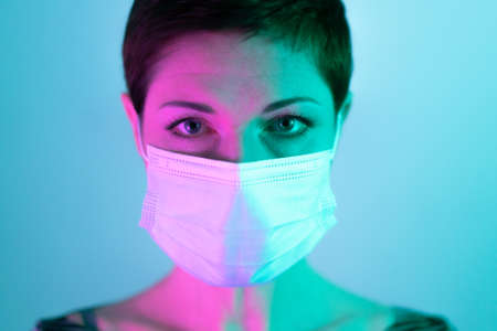 Beautiful woman with short hair wearing a face mask, looking worried and depressed in violet and green lighting. Life in quarantine. Dealing with coronavirus restrictions. Life during pandemic.の写真素材