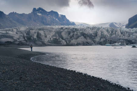 Tourist walks by glacier lagoon on a cold, cloudy day in southern Iceland. Glacier falls into the water. Harsh, dramatic, misty weather.の写真素材