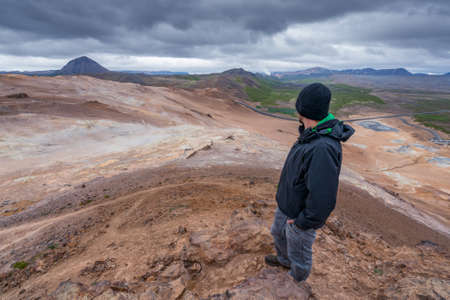 A man overlooking volcanic Icelandic landscape from the top of Namafjall. Dramatic clouds in the sky. Road in the distance winds through the land. Hverir, boiling mud area.の写真素材
