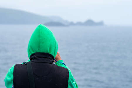 Man in a green hoodie takes a photo of a remote island in the vast waters of northern Atlantic....