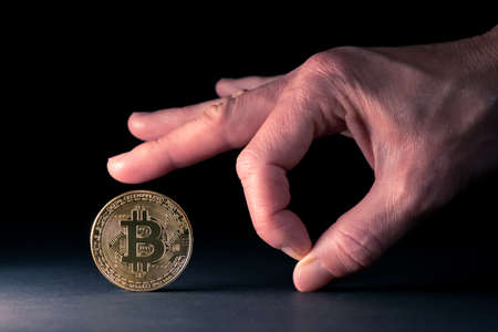 Concept for bitcoin crisis, blockchain bans and restrictions. Hand with fingers ready to send the golden bitocin away from a illuminated surface on black backgroundの写真素材