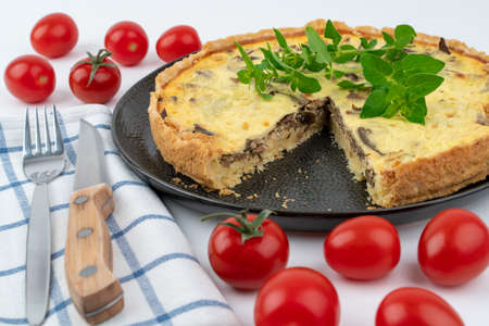 Closeup wide studio shot of freshly baked yellow French salty cake, or quiche with mushrooms, red cherry tomatoes, green mint and kitchen cloth, on black design plate on white background. Cutlery set.の写真素材