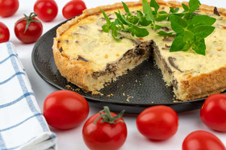 Closeup wide studio shot of freshly baked yellow French salty cake, or quiche with mushrooms, red cherry tomatoes, green mint and kitchen cloth, on a black design plate, on white backgroundの写真素材