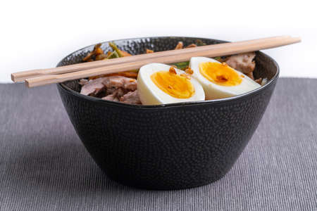 Studio shot of black bowl of ramen noodles with boiled eggs, veggies and turkey meet, ready to be poured with boiling broth. Wooden chopsticks. Freshly prepared and good looking Asian food.の写真素材