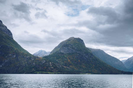 Majestic mountains tower above lake in northern Norway on a cloudy day of Arctic summer. Small houses between the mountains far in the distance. Mysterious Norwegian landscape.の写真素材