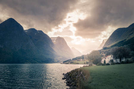 Stormclouds with sun shining through them above dramatic mountain range of Norwegian fjord. Viewed from the shore with white Scandinavian house nearbyの写真素材