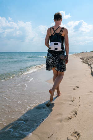 Hel, Poland - 08.01.2021: Vertical shot of a woman in black and white skirt, with a small bag, walking barefoot away from the viewer on a beach. Beautiful summer morning by the Baltic sea.のeditorial素材