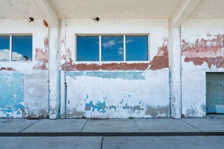 Detail shot of cracked white, blue and red wall of a port warehouse with blue sky reflecting in its long horizontal windows. Old building decay.の写真素材