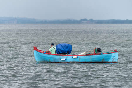 Fisherman in green t-shirt smoking a cigarette sits in front of his small blue boat, sailing into Baltic sea near Jastarnia, Poland, on a cloudy summer day. Land in the haze on distant horizon.の写真素材