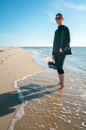 Vertical shot of a woman in dark outdoor clothes with shoes in her right hand, standing barefoot on a beach, with tide washing her feet. Beautiful summer morning by the Baltic sea. Seaside walk.の写真素材