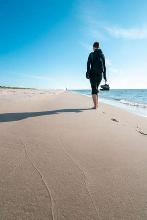 Vertical shot of a woman in dark outdoor clothes with shoes in her right hand, walking barefoot away from the viewer on a beach. Beautiful summer morning by the Baltic sea. Summertime seaside walk.の写真素材