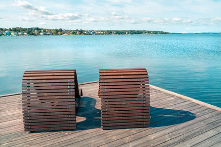 Two wooden beach beds or chairs on a wooden pier next to the sea with cityscape in the background. Sunny summer day by the Baltic Sea in Vastervik, southern Sweden. Summer vacation by the sea.の写真素材