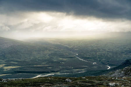 Bright sunlight comes through thick clouds and illuminates heavy rainfall in the distance above remote rough arctic valley. Dramatic weather in Rapadalen, Sarek National park, Swedish Lapland.の写真素材