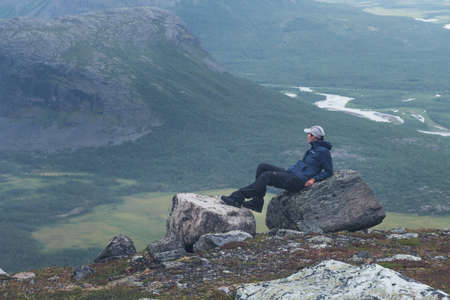 Female hiker in blue jacket overlooking remote arctic valley on a cloudy day of arctic summer. Hiking in Sarek National Park, Sweden, with Rapa River valley bellow. Freedom and adventure.の写真素材