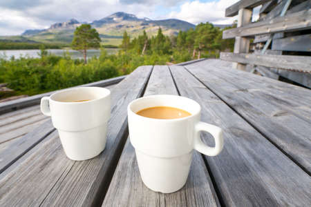 Two white cups filled with coffee with milk on a wooden table with arctic landscape of Swedish Lapland in the backgroundの写真素材