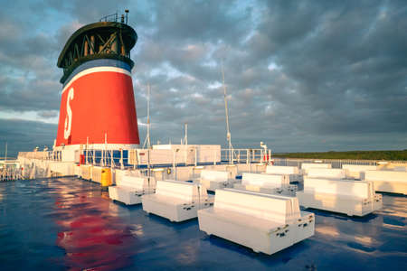 Karlskrona, Sweden - 09.14.2021: Top deck of Stena Line Spirit ship heading for Sweden in nice autumn morning. Big red chimney and white benches on wet blue deck.のeditorial素材