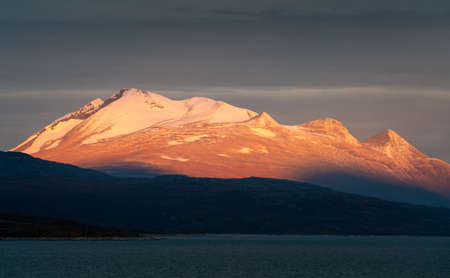 Epic sunrise light illuminating rocky, glacier covered peak with dark cloudy sky in the background....