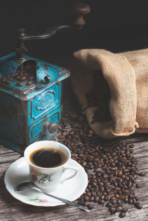 Vertical studio shot of still life with vintage manual green metal coffee grinder, jute bag and coffee beans on old wooden table. Vintage white cup of hot coffee with steam going up. Coffee concept.の写真素材