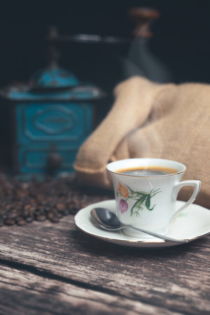 Vertical studio shot of still life with vintage manual green metal coffee grinder, jute bag and coffee beans on old wooden table. Vintage white cup of hot coffee with steam going up. Coffee concept.の写真素材