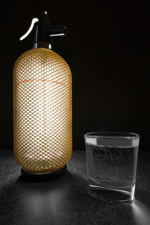 Vertical wide studio shot of backlit, luxury looking vintage soda siphon with golden color case on stone desk. Glass of soda.の写真素材